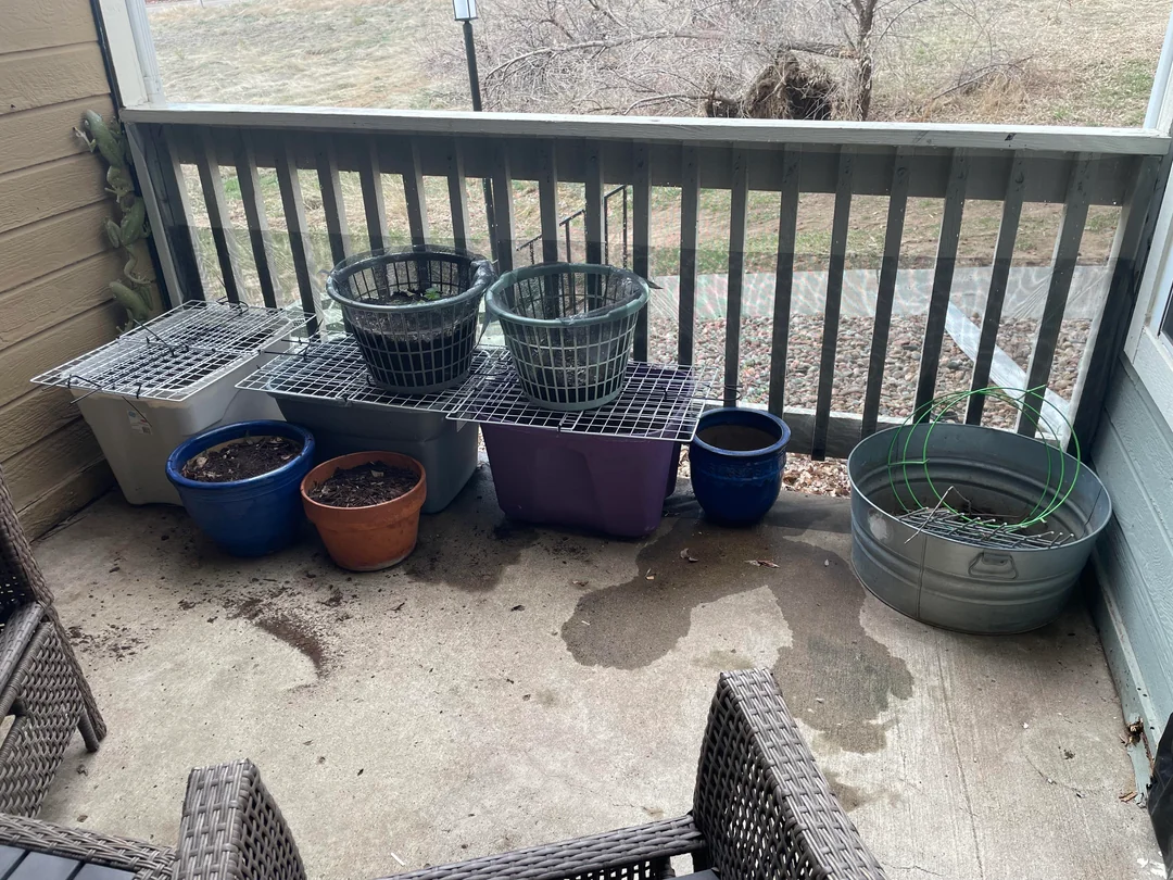 Several empty and partially filled plant pots and containers are arranged on a small outdoor balcony with a concrete floor, overlooking a grassy area. Two patio chairs are visible in the foreground.