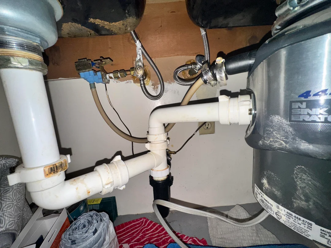 Under-sink plumbing setup showing white PVC pipes, hose connections, a garbage disposal unit on the right, and water valves and electrical outlet mounted on the back wall. Various supplies are visible on the floor.