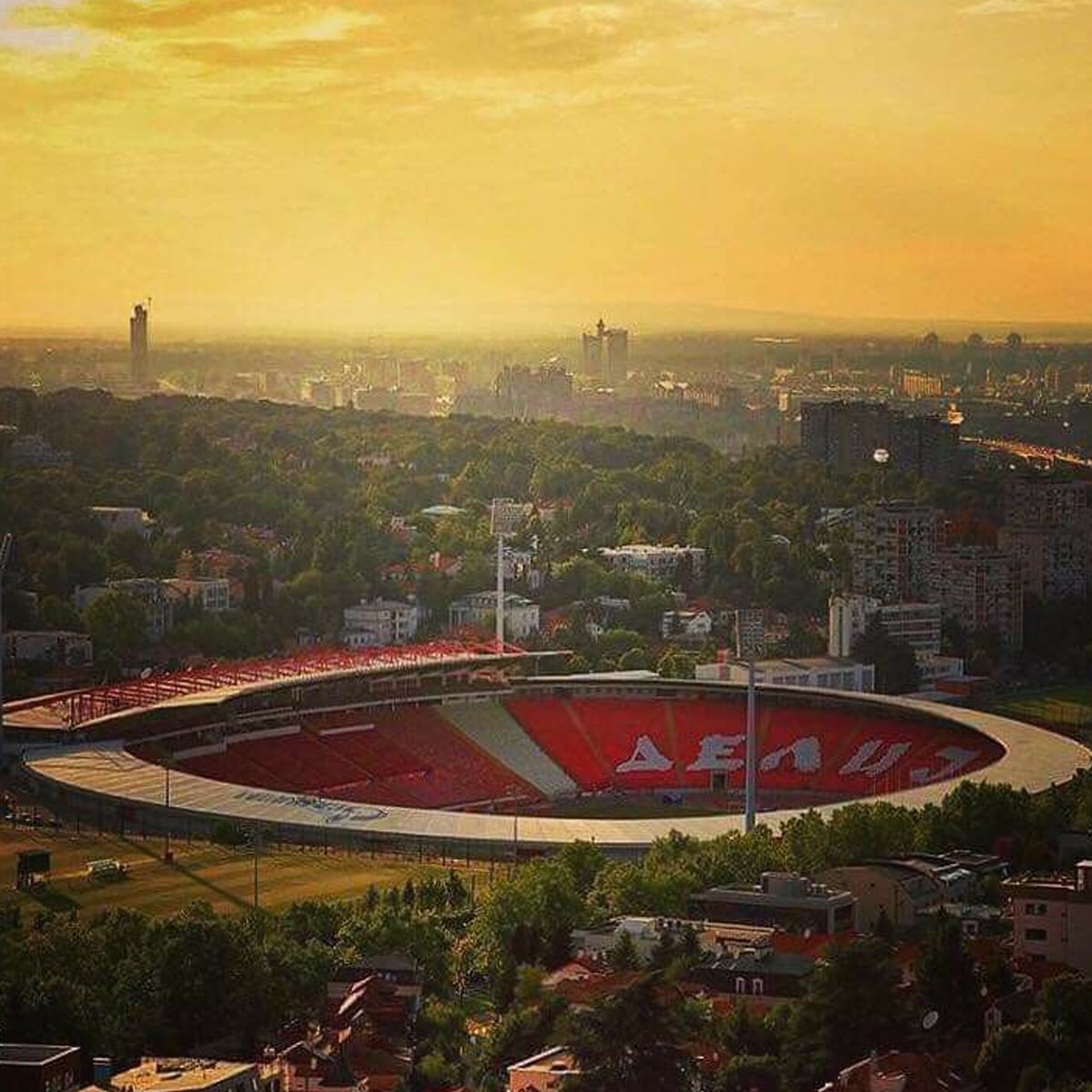 A large outdoor stadium with red and white seats, surrounded by trees and city buildings, is seen at sunset under a golden sky.
