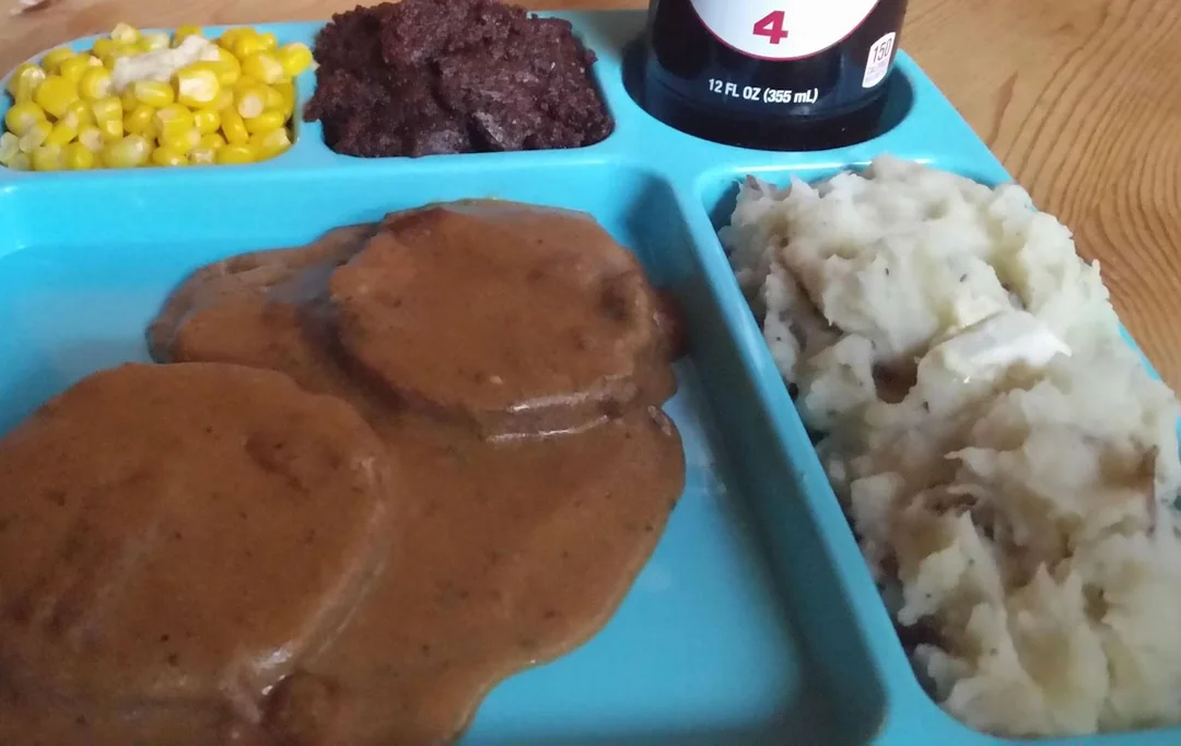 A blue cafeteria tray with mashed potatoes, corn, chocolate brownie, and Salisbury steak patties covered in brown gravy. A can of soda is partially visible in the background.