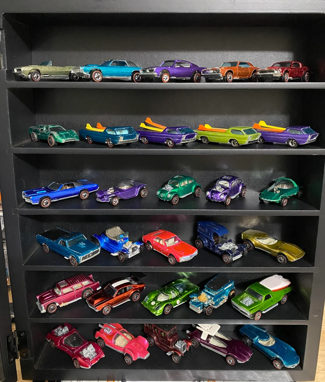 A black display shelf holding five rows of colorful toy cars, mostly vintage Hot Wheels and Matchbox models, in various styles and bright colors, neatly arranged across each shelf.