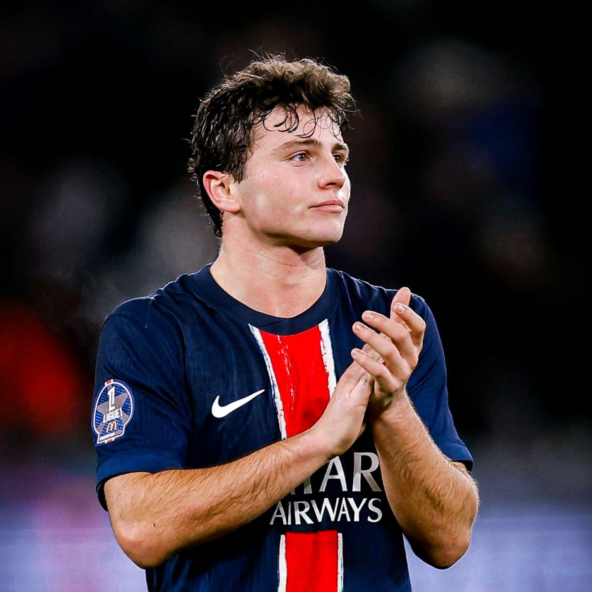 A soccer player wearing a navy blue Paris Saint-Germain jersey with red and white stripes stands on the field, looking to the side and clapping with both hands. The background is blurred.