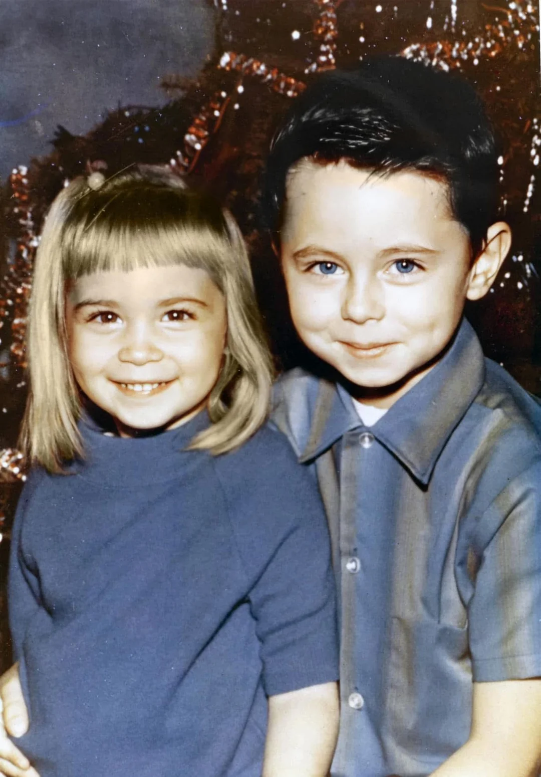 A young girl with blonde hair and a blue dress smiles beside an older boy with dark hair and a blue shirt, both posing in front of a decorated Christmas tree.