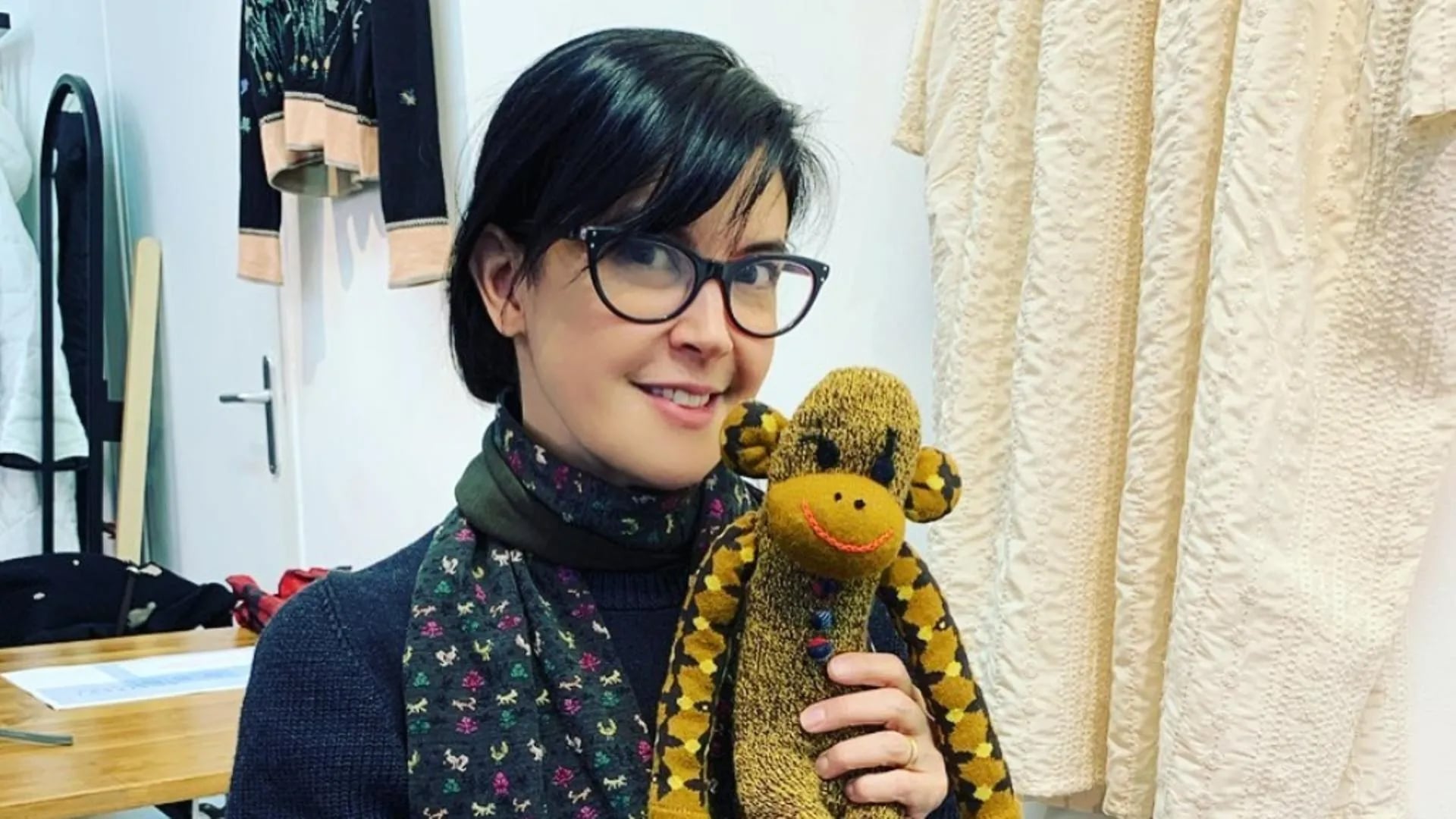 A woman wearing glasses and a patterned scarf smiles while holding a brown and yellow stuffed monkey with a red smile, standing in a room with light-colored clothing hanging in the background.
