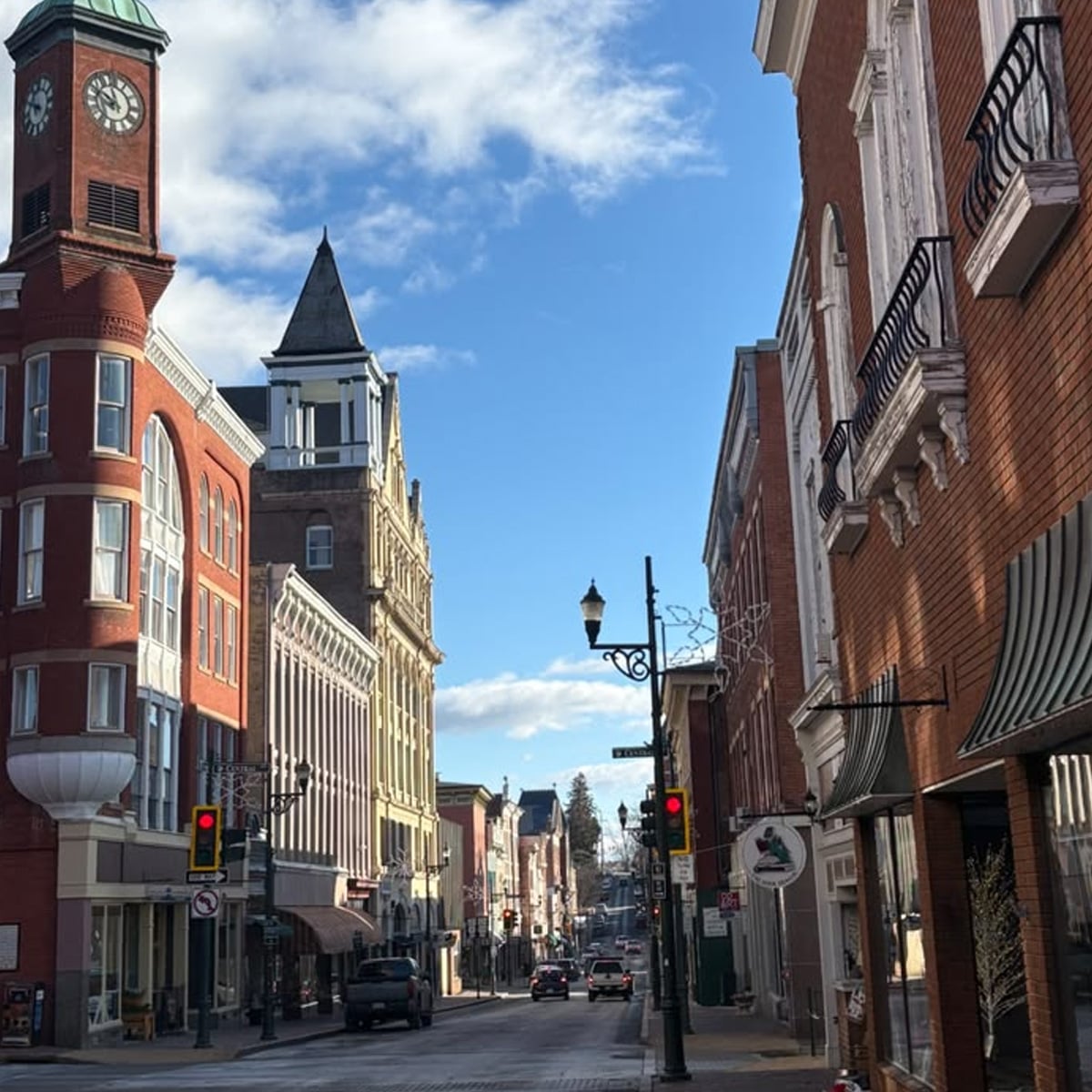 A street in a small town with historic brick buildings, a clock tower on the left, storefronts, cars on the road, and blue sky with scattered clouds.