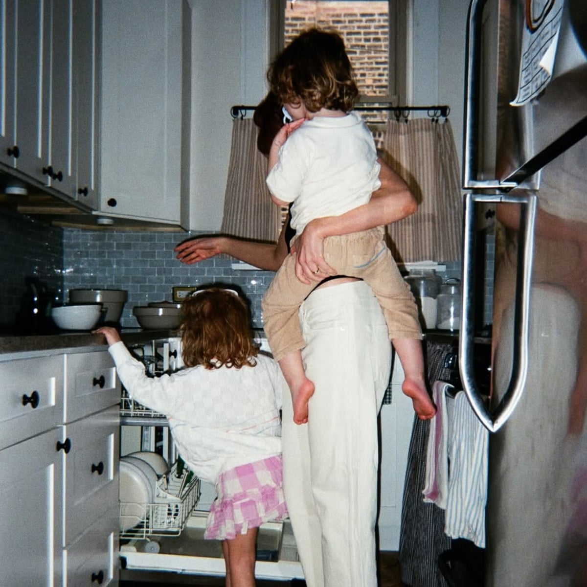 An adult stands in a kitchen holding a child on their hip while another child helps load the dishwasher. The kitchen has white cabinets, a window, and dishes on the counter.