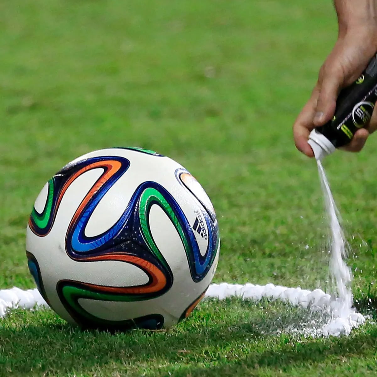 A soccer referee sprays white foam on the grass to mark a line beside a colorful soccer ball, preparing for a free kick during a match.