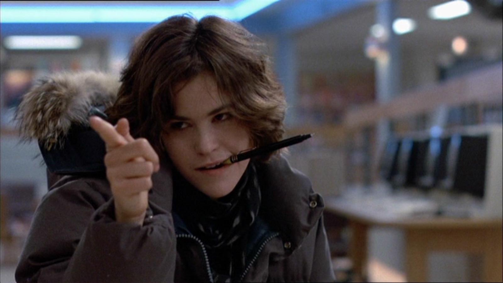A person with short brown hair wearing a hooded winter coat holds a pen in their mouth and points playfully with their finger inside a room with computers and fluorescent lighting.