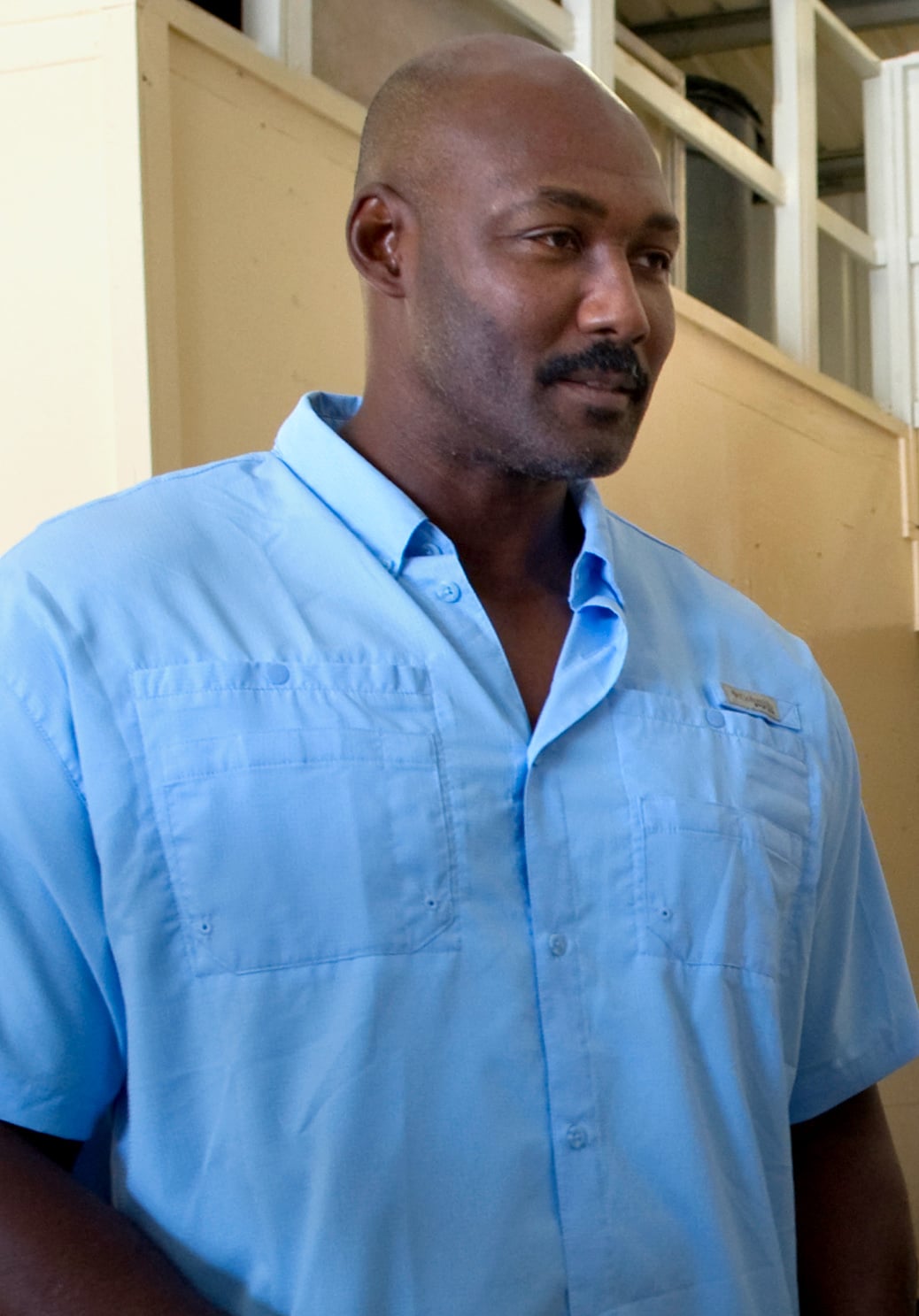 A man with a shaved head and mustache is wearing a light blue button-up shirt, standing indoors in front of a beige wall and partition.