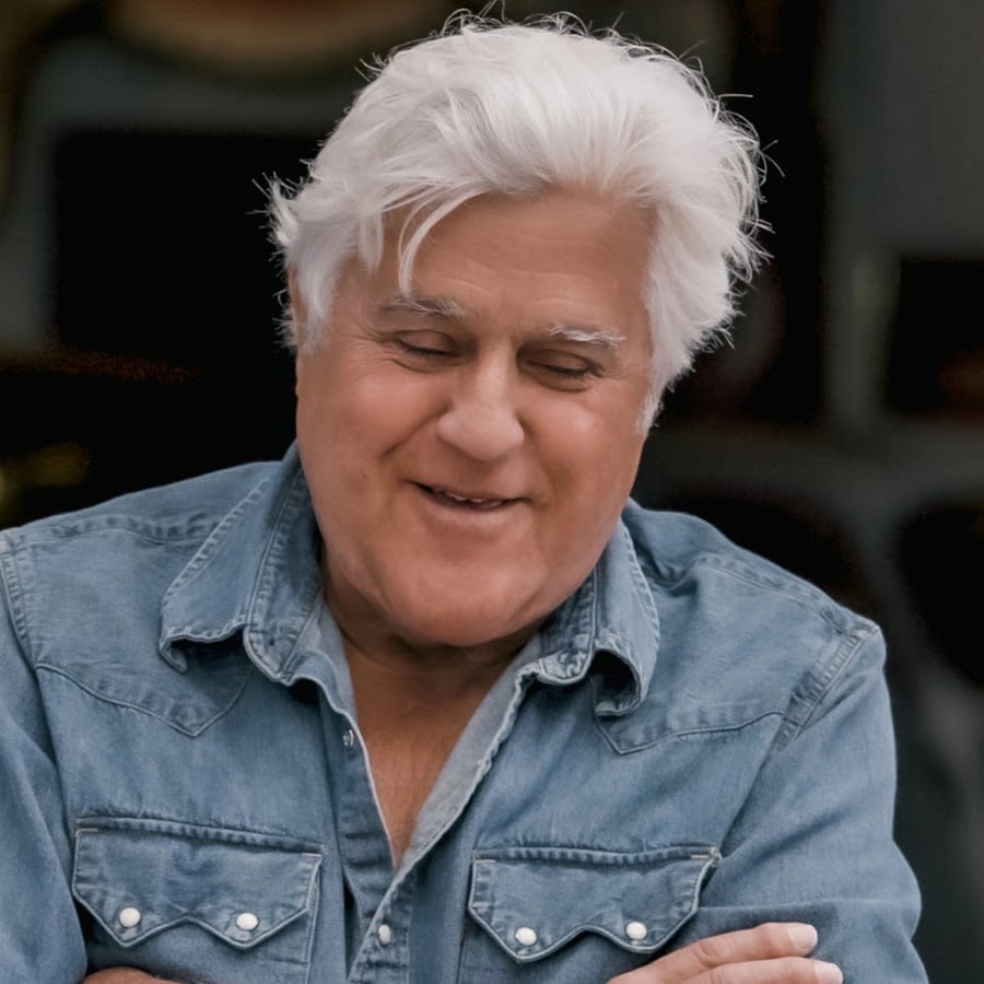 An older man with white hair smiles with his arms crossed, wearing a light denim button-up shirt. The background is blurred and dark, focusing attention on his relaxed expression.