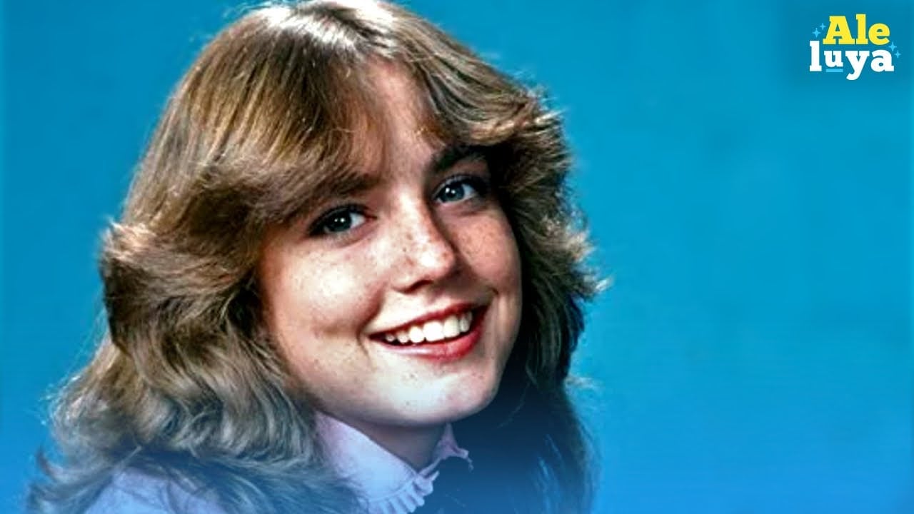 A teenage girl with light brown, wavy hair and freckles smiles at the camera against a blue background. She is wearing a light pink, ruffled-collar shirt. The word "Aleluya" is in the top right corner.