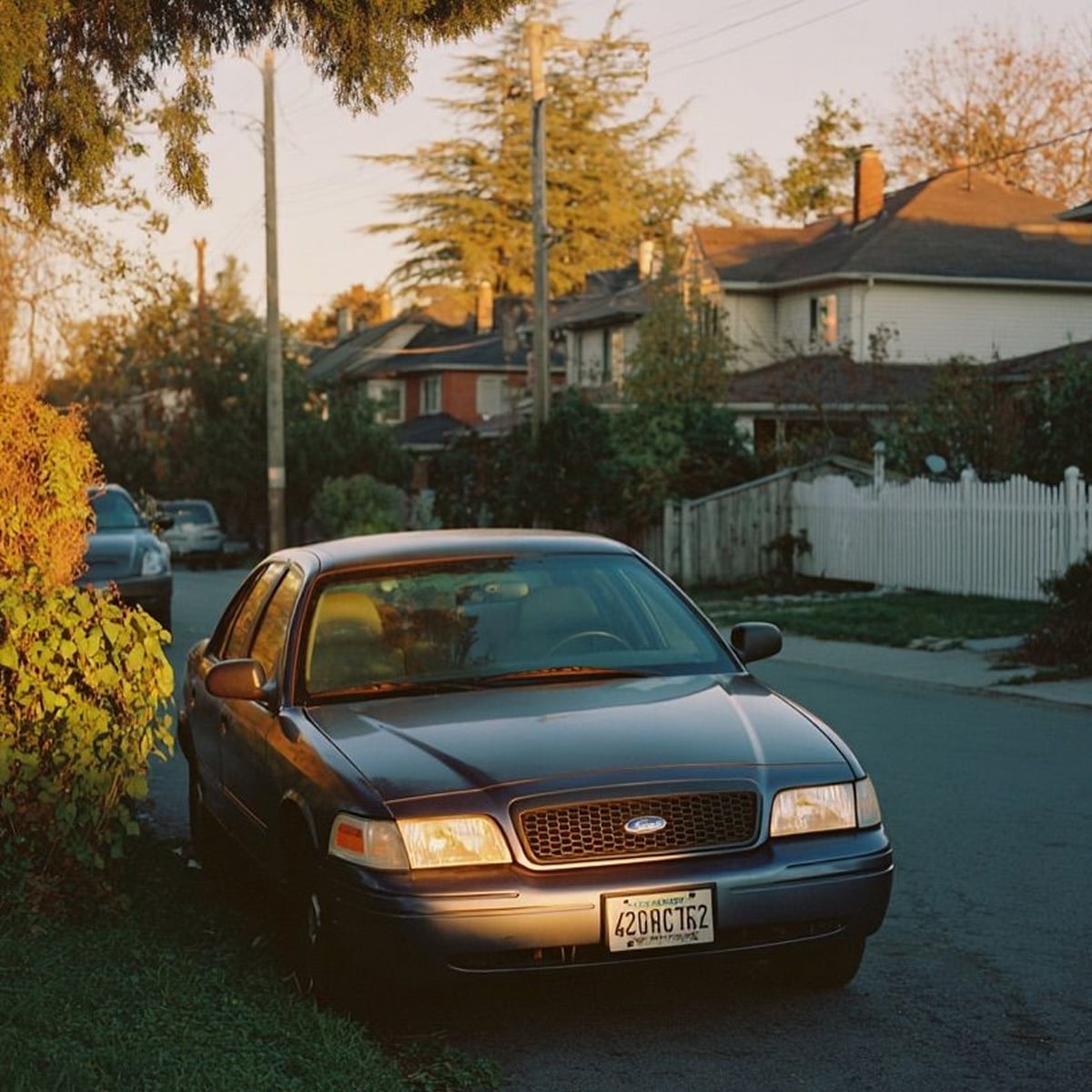 A parked dark-colored Ford sedan sits on a residential street at sunset, surrounded by greenery and houses with white picket fences. Sunlight casts a warm glow over the scene.
