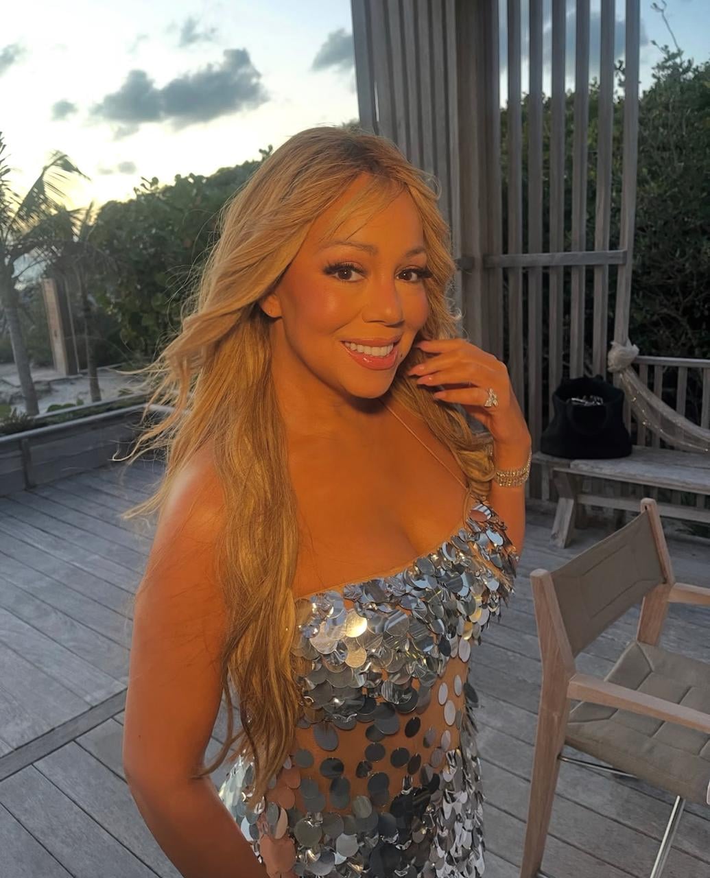 A woman with long, wavy blonde hair smiles outdoors at sunset, wearing a strapless dress adorned with large silver sequins. She stands on a wooden deck surrounded by greenery and outdoor furniture.