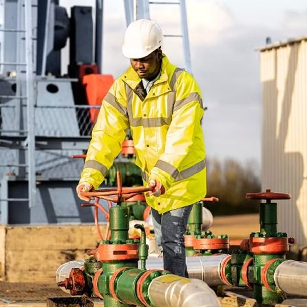 A worker wearing a yellow high-visibility jacket and white hard hat operates a large red valve on industrial piping at an outdoor facility.