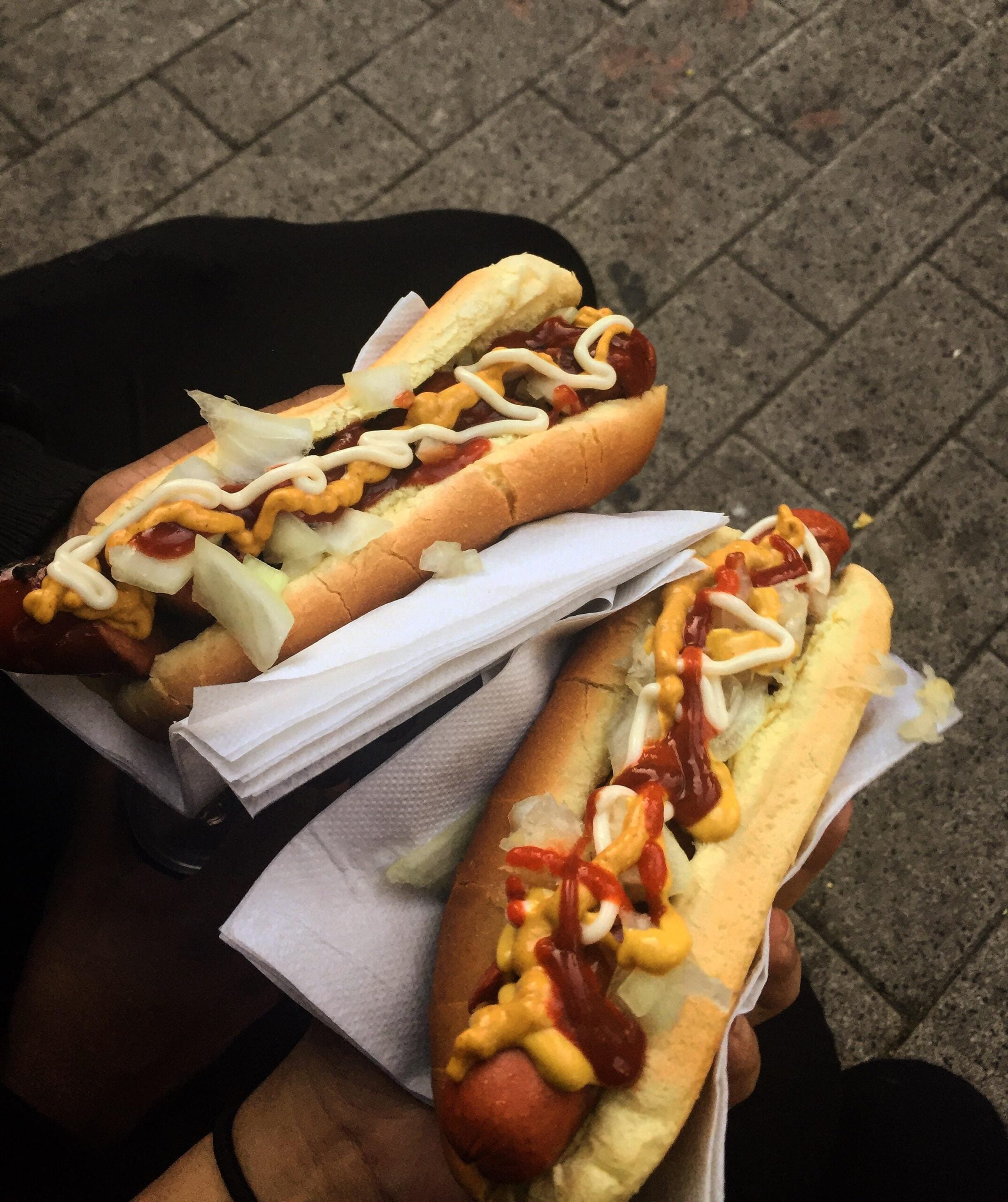 Two hands holding hot dogs in buns, topped with chopped onions, ketchup, mustard, and mayonnaise, placed on white napkins. The background shows a gray pavement.