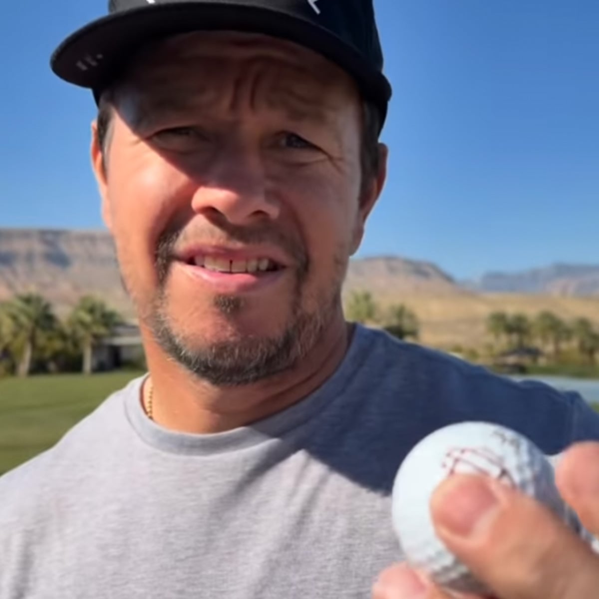 A man wearing a black cap and gray shirt holds a golf ball up to the camera on a sunny day at a golf course, with mountains and palm trees in the background.