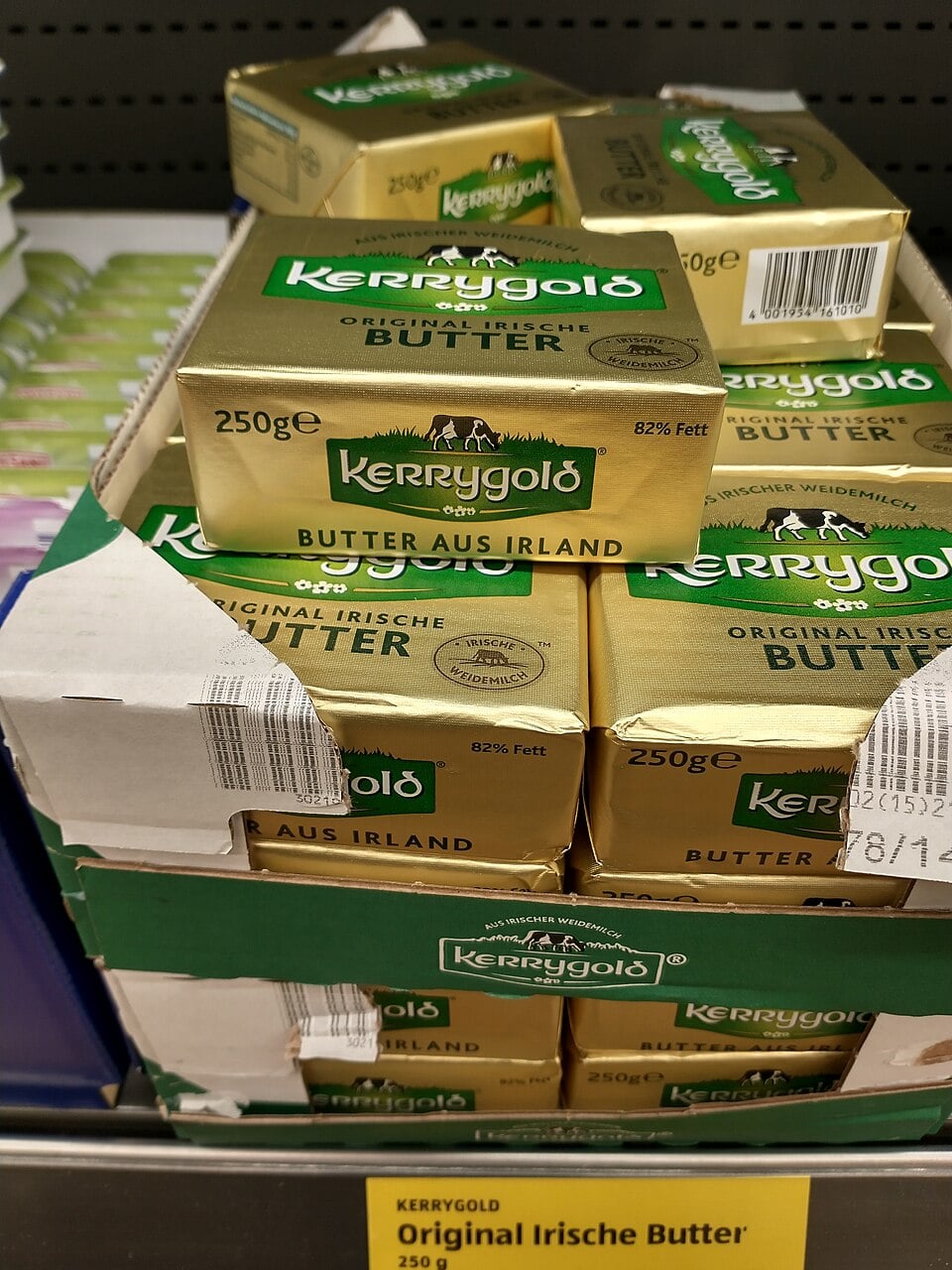 Several packs of Kerrygold Irish butter, wrapped in gold foil with green labels, are stacked in a display box on a grocery store shelf. The packs are labeled "250g" and "Butter aus Irland.