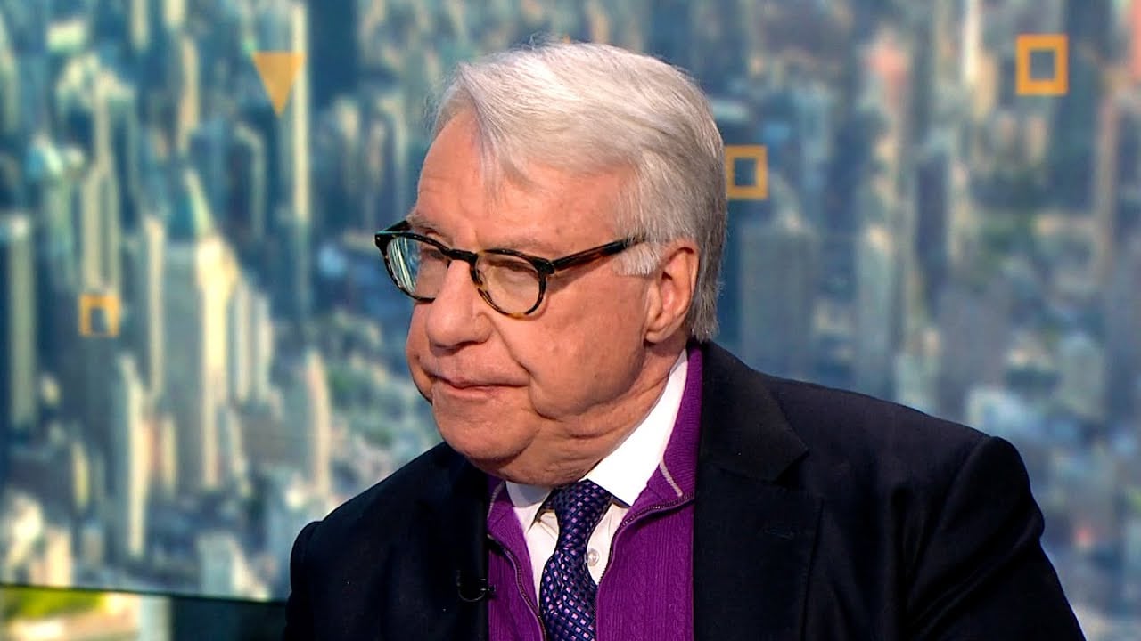 An older man with white hair, glasses, and a dark suit over a purple sweater sits in front of a blurred cityscape background, looking slightly to the side with a serious expression.