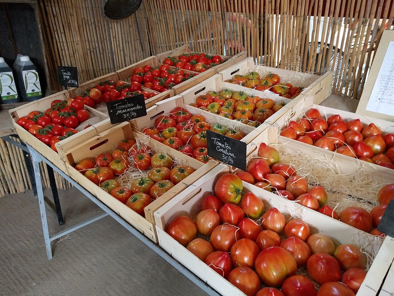 Wooden crates filled with various kinds of tomatoes are displayed on a table at a market stall. Small chalkboard signs label the types of tomatoes and their prices. Bottles and wooden paneling are visible in the background.