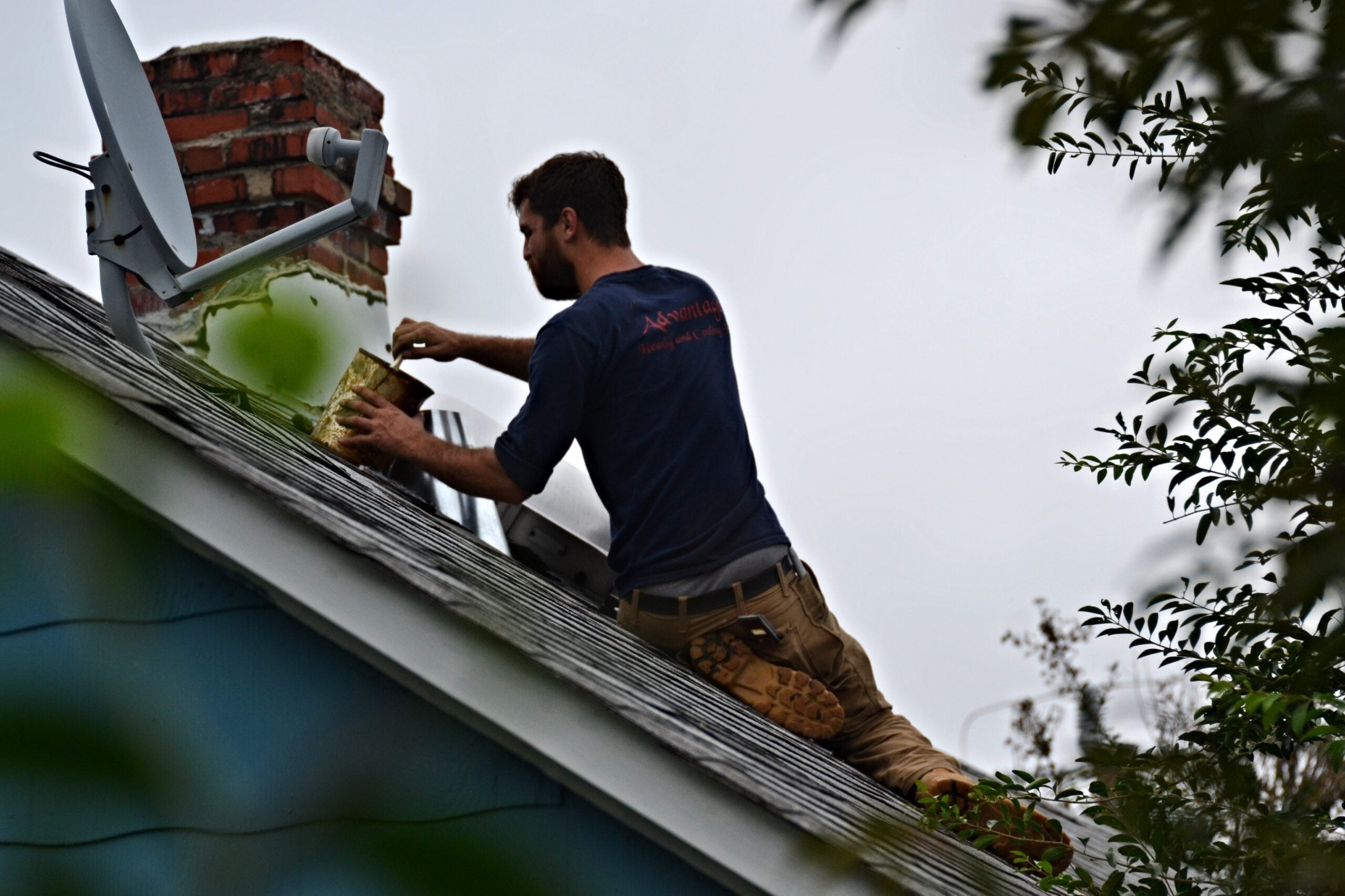A person kneels on a sloped roof, painting near a chimney and satellite dish. Trees frame the scene, and the sky is overcast. The individual wears a tool belt and a dark long-sleeve shirt.