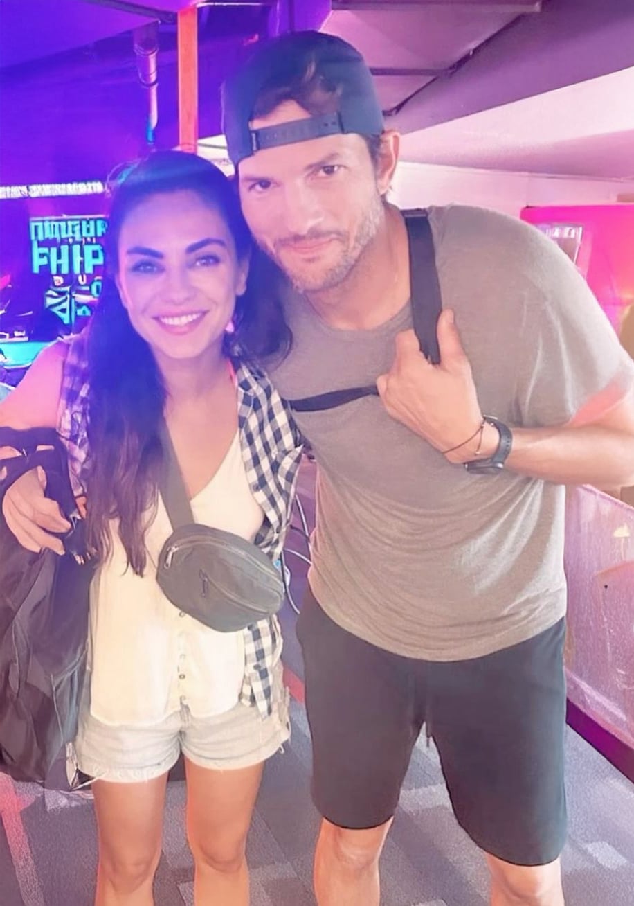 A woman in a sleeveless plaid shirt and shorts stands next to a man in a t-shirt, shorts, and a backwards cap. They are both smiling in an indoor setting with colorful lights and arcade machines in the background.