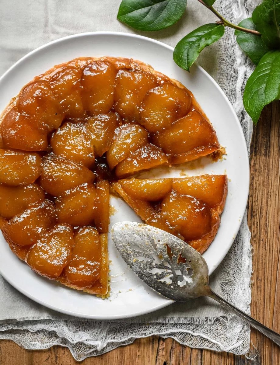 A round tarte Tatin with glossy caramelized apples sits on a white plate, one slice missing. A vintage pie server rests on the side, and a lace-edged napkin and green leaves decorate the rustic wooden table.