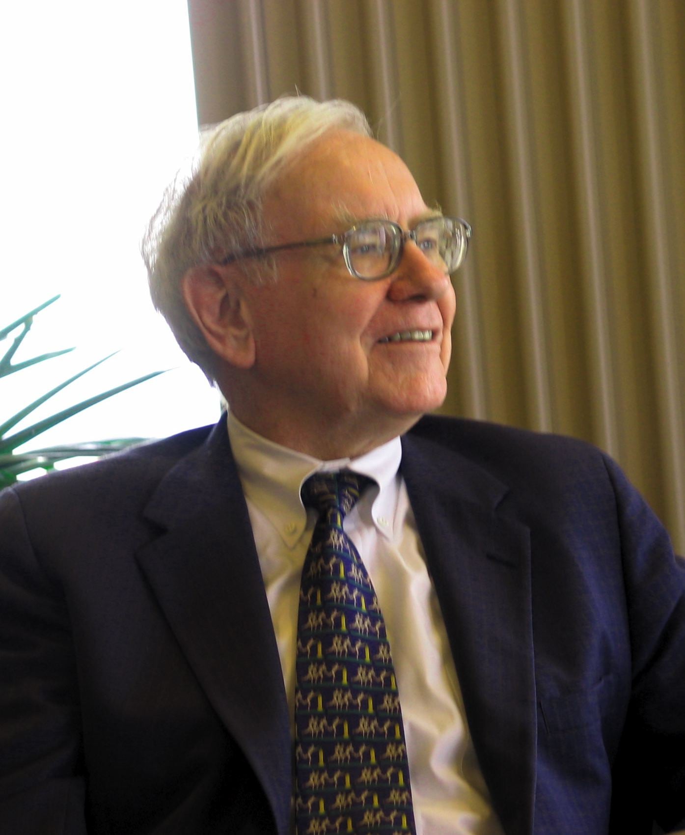 An older man with white hair and glasses, wearing a dark suit, white shirt, and patterned tie, is sitting and smiling while looking to his left. Vertical blinds and a plant are visible in the background.