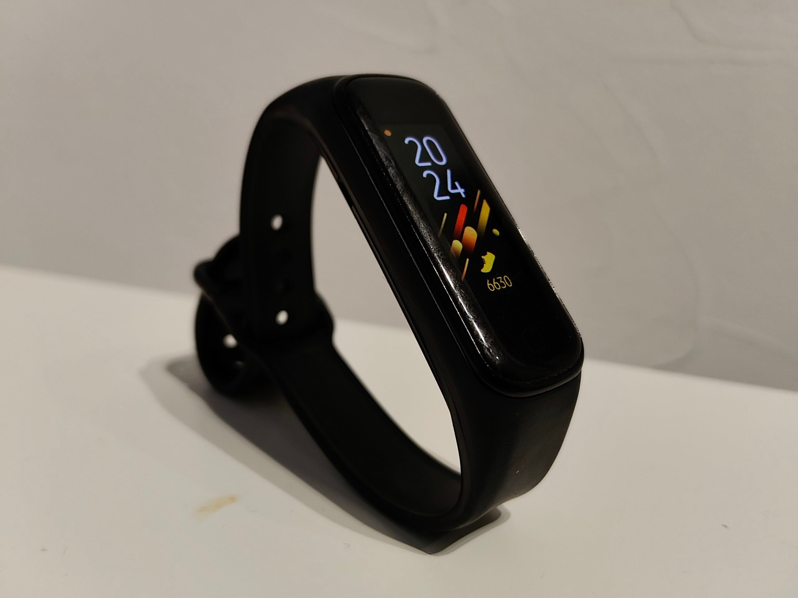 A black fitness tracker with a rectangular screen displays the time "20:24", a step count, and colorful activity icons, resting on a white surface against a light background.