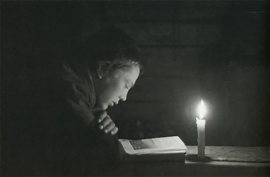 A person sits at a table reading a book by candlelight in a dimly lit room, with the candle casting a warm glow on their face and the book.