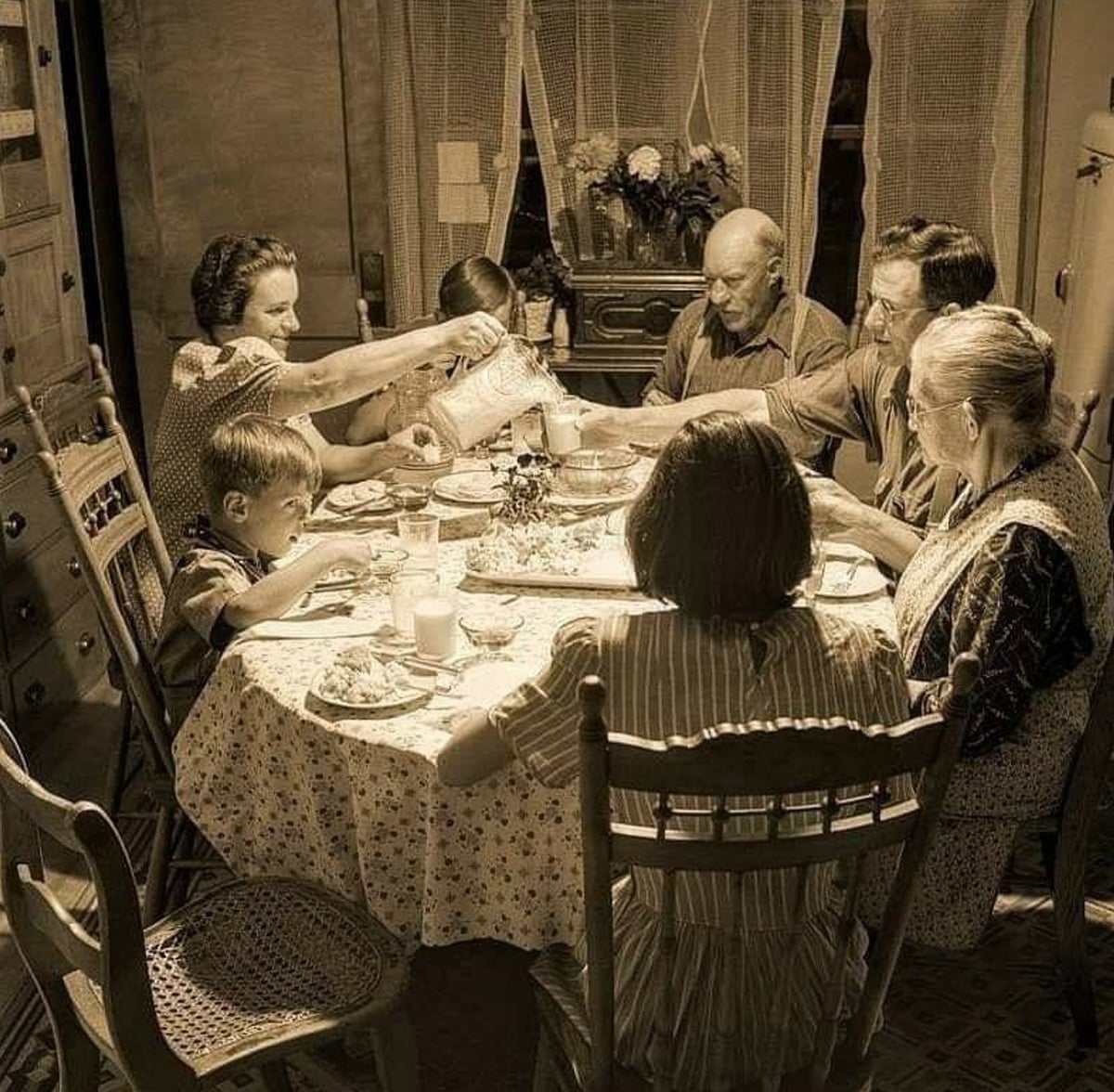 A family of seven, including children and elderly adults, sits around a dining table sharing a meal in a vintage-style kitchen, with a woman pouring milk. The atmosphere is warm and nostalgic.