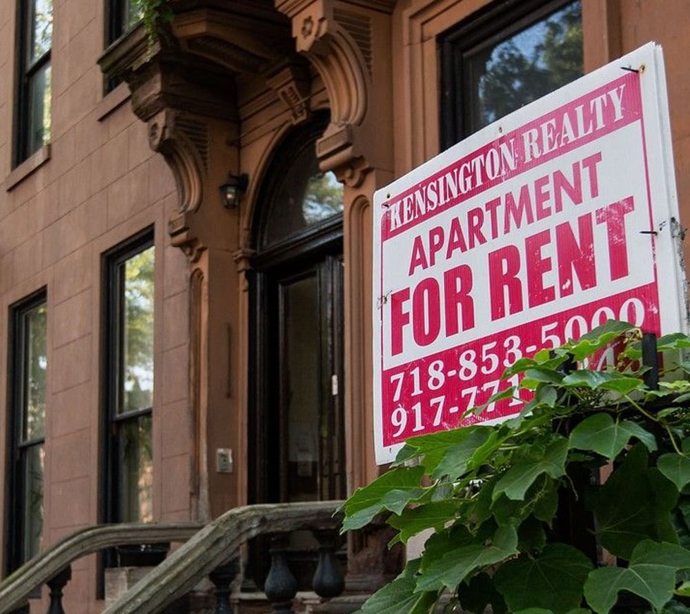 A red and white "For Rent" sign for Kensington Realty stands in front of a brownstone apartment building with large windows and a black doorway, partially surrounded by green plants.