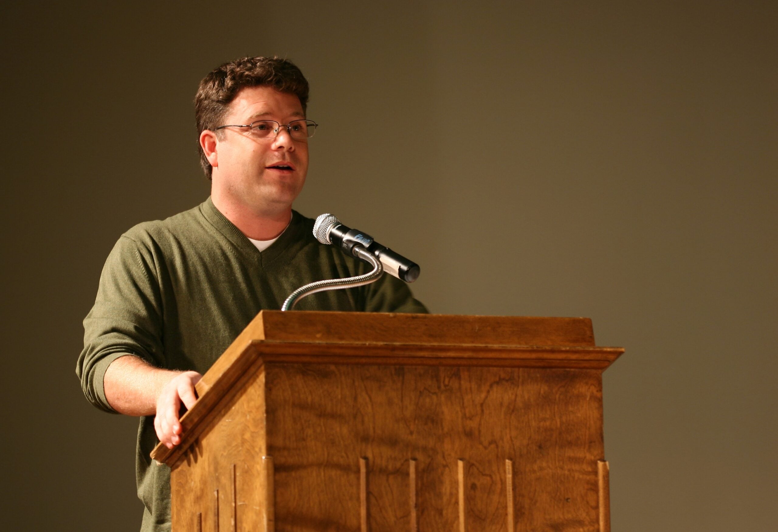 A man wearing glasses and a green sweater stands at a wooden podium speaking into a microphone with a neutral background.