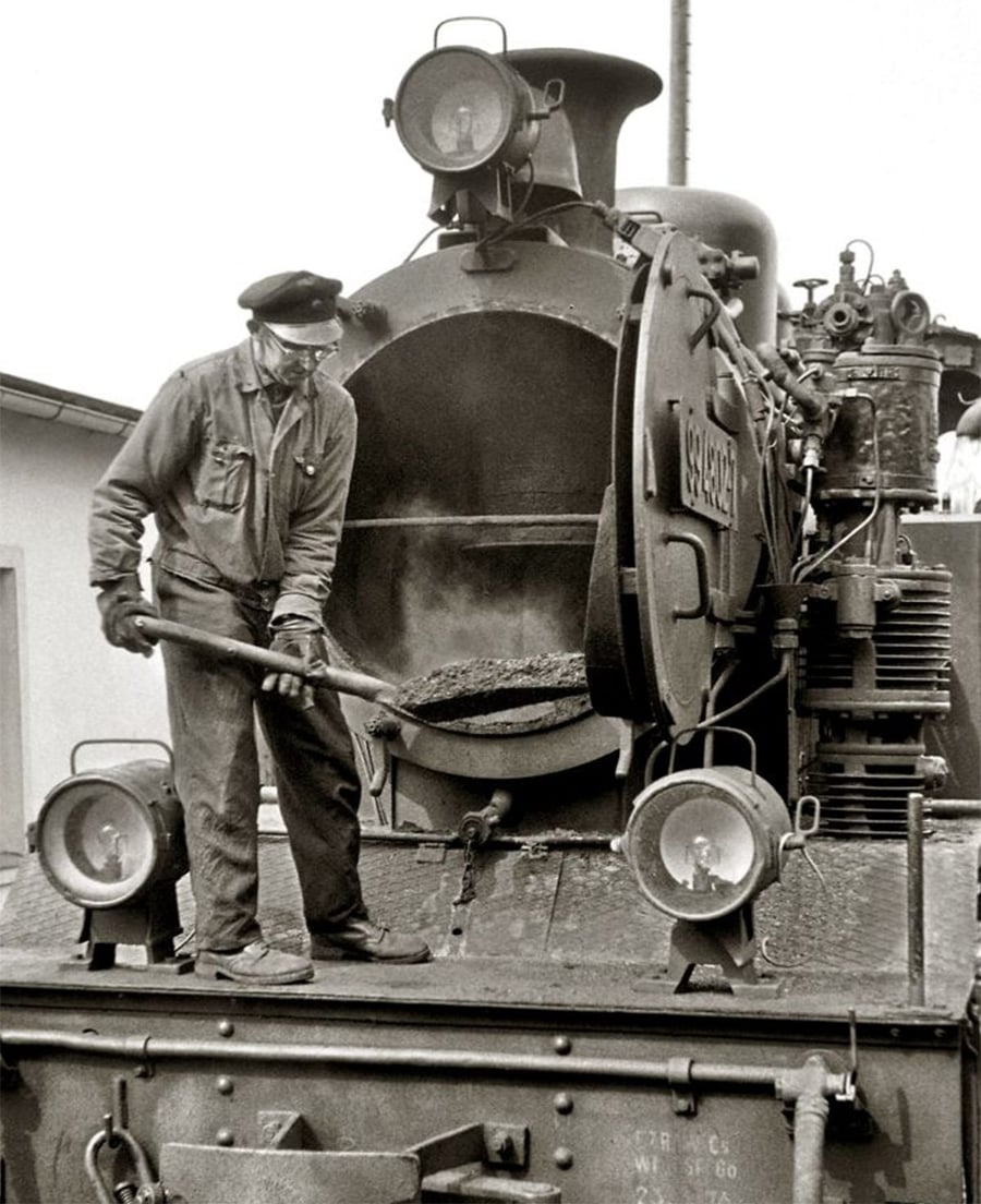 A worker in uniform and cap shovels coal into the open firebox of a steam locomotive. The scene is black and white, and the worker stands on the front platform of the train engine.