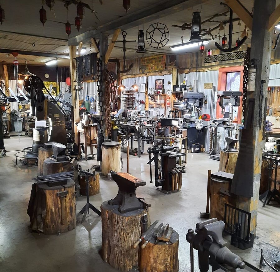 Spacious blacksmith workshop with multiple anvils on tree stumps, tools, equipment, and workbenches. The shop has a rustic, organized appearance with machinery and metalworking tools throughout.