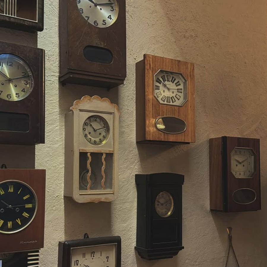 A collection of vintage wall clocks with different shapes, sizes, and designs hangs on a beige textured wall. Most of the clocks are wooden and display various times.