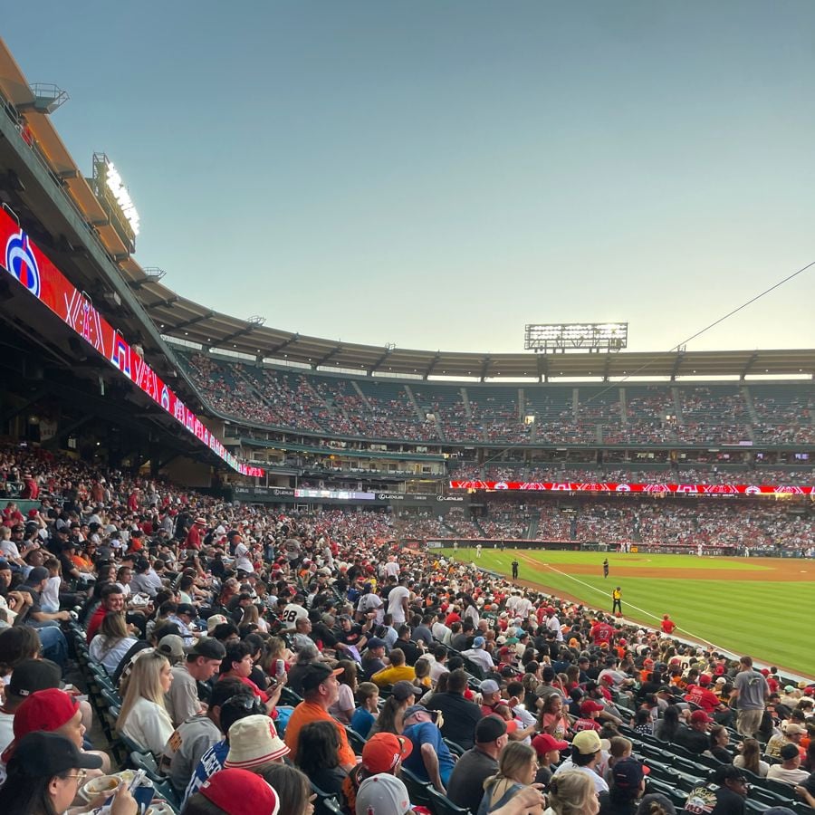 A large crowd fills the stands at a baseball stadium during a game at sunset. Fans watch the field intently, with the scoreboard and stadium lights visible in the background.