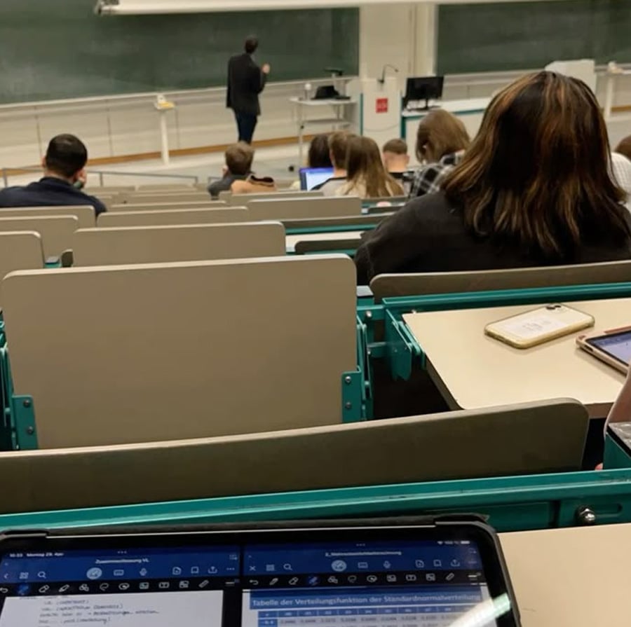 A university lecture hall with students seated facing a chalkboard. Some students use laptops and tablets, while the lecturer stands at the front near the board, writing or presenting.