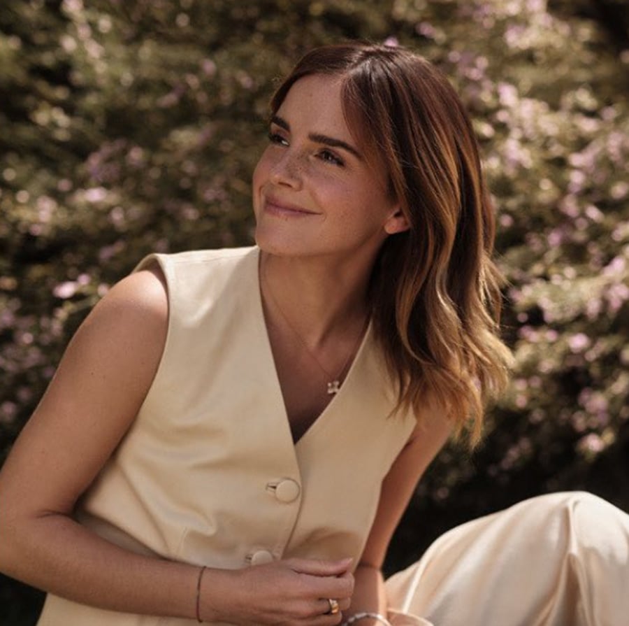A woman with shoulder-length brown hair, wearing a sleeveless beige top, smiles softly while sitting outdoors in front of blurred greenery.