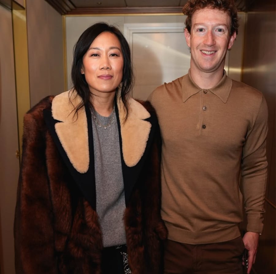 Two people stand side by side indoors. The woman wears a large fur coat with a beige collar over a gray sweater, and the man wears a tan long-sleeve polo shirt and brown pants. Both are smiling.