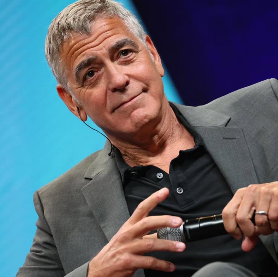 A man with short gray hair in a gray suit and black shirt sits holding a microphone, looking ahead with a slight smile, against a blue and purple background.