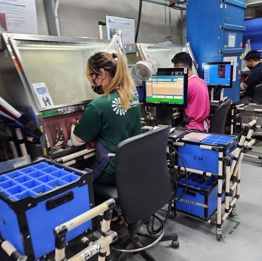 Three people sit at workstations with monitors and blue storage bins, performing manual tasks inside enclosed boxes using built-in gloves at an industrial or manufacturing facility.