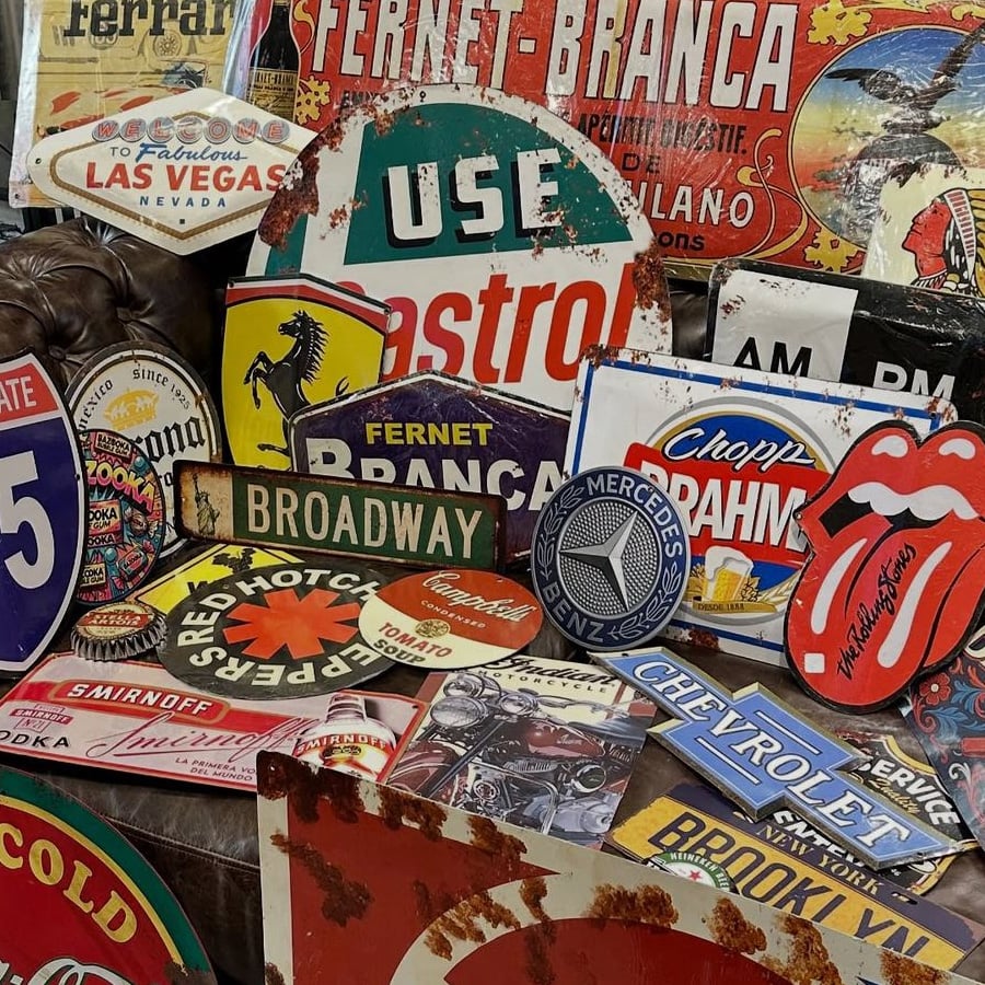 A cluttered display of vintage metal signs, including brands like Chevrolet, Mercedes-Benz, Fernet-Branca, and Castro, plus street signs, a Rolling Stones logo, and other retro advertisements.