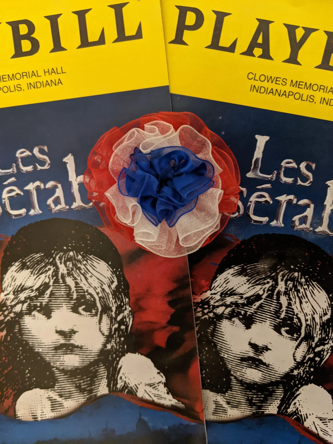 Two Playbills for "Les Misérables" at Clowes Memorial Hall in Indianapolis are shown, partially overlapping. A red, white, and blue fabric flower sits on top, matching the colors of the French flag.