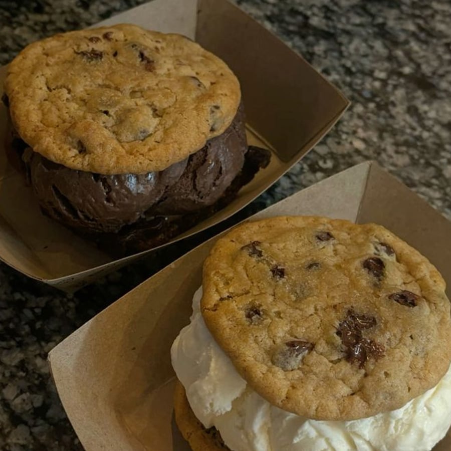Two ice cream sandwiches in brown paper trays, each made with two chocolate chip cookies. One has chocolate ice cream, the other has vanilla ice cream, and both are placed on a granite countertop.