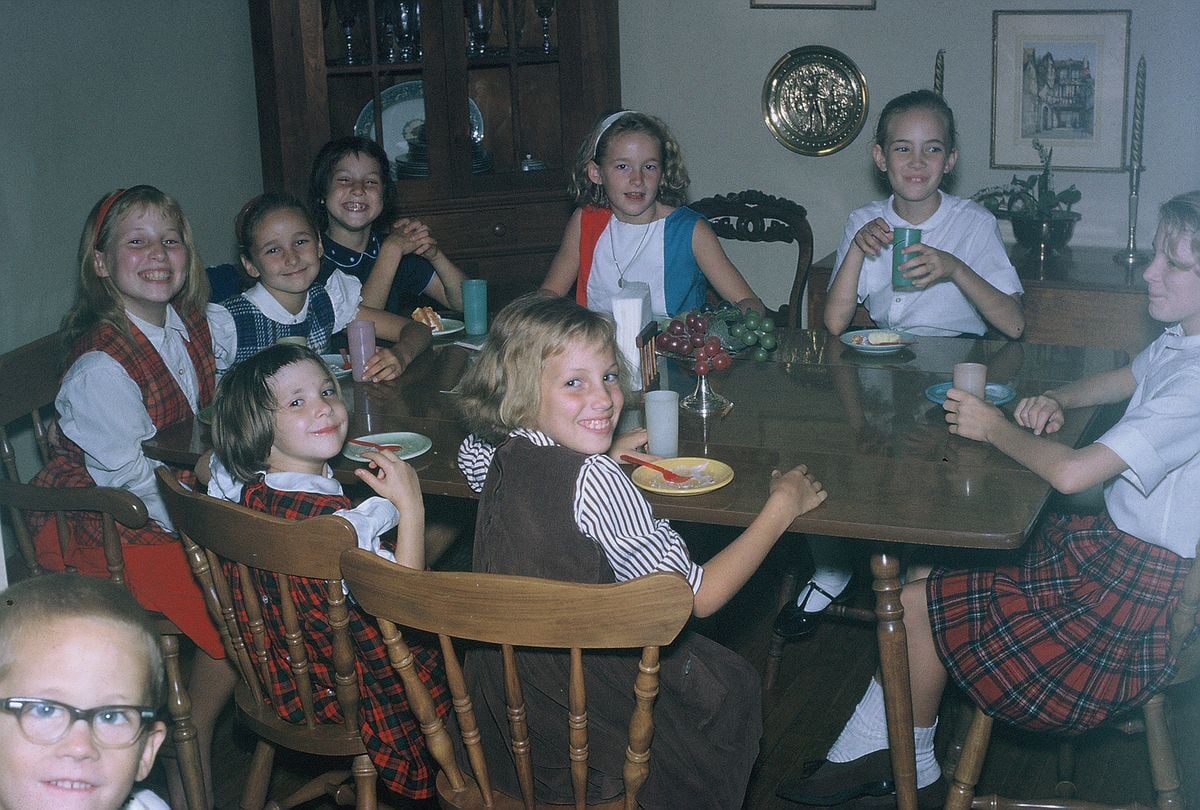 Eight young girls, dressed in 1960s-style clothing, sit around a wooden dining table with dishes, glasses, and food. They are smiling and appear to be enjoying a gathering in a cozy, well-decorated room.