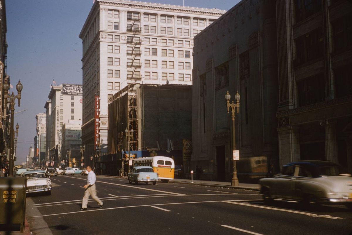 A man crosses a wide city street with vintage cars and a yellow bus. Tall buildings line both sides, and the sunlight casts long shadows. The scene appears to be from the mid-20th century.