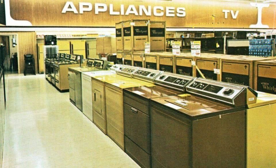 A vintage store interior with rows of washing machines, dryers, and other appliances in brown, gold, and white colors, beneath a large "APPLIANCES TV" sign. Cardboard boxes are stacked in the background.