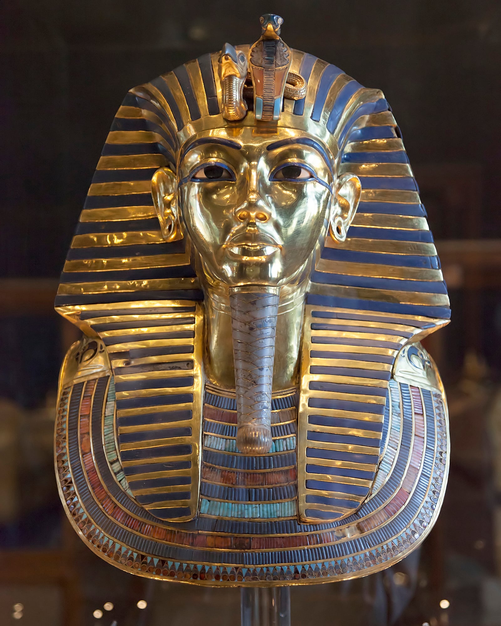 A highly detailed, gold and blue striped funerary mask of an ancient Egyptian pharaoh, featuring a false beard, elaborate collar, and royal headdress with a cobra and vulture on the forehead.