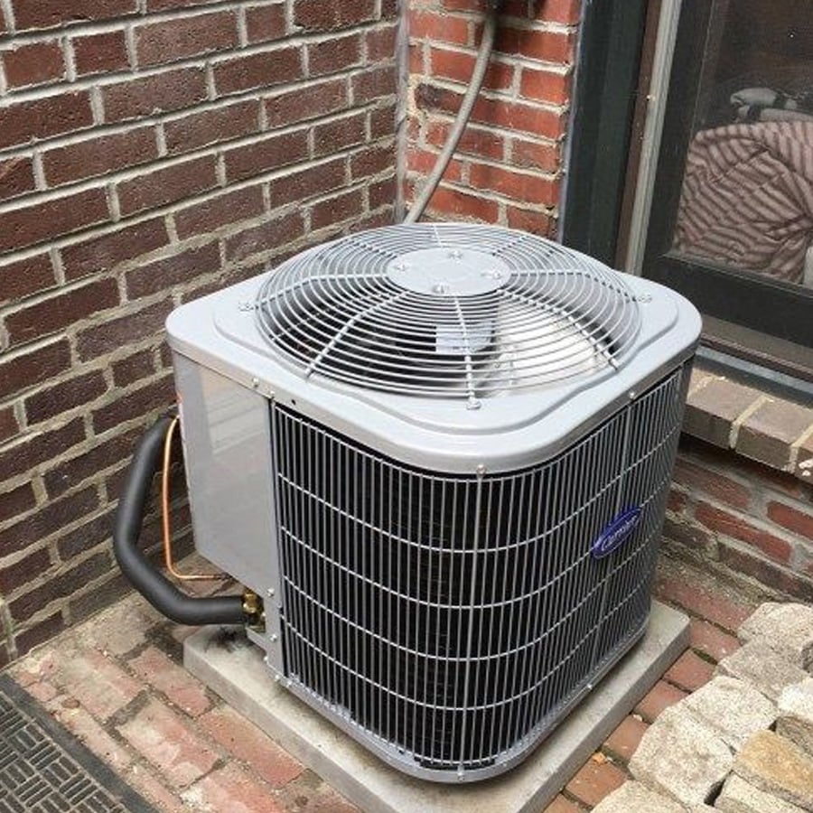 A gray outdoor air conditioning unit sits on a concrete pad next to a brick wall and window, with electrical wiring connected to the building.