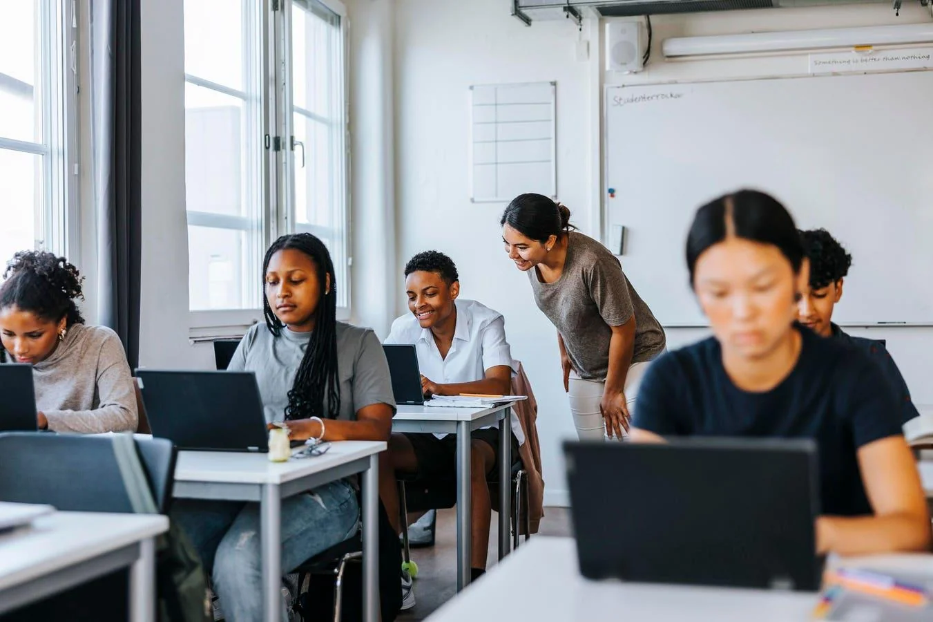 A teacher leans over to assist a student in a classroom where several students are working on laptops at their desks. The room is bright with large windows and a whiteboard in the background.